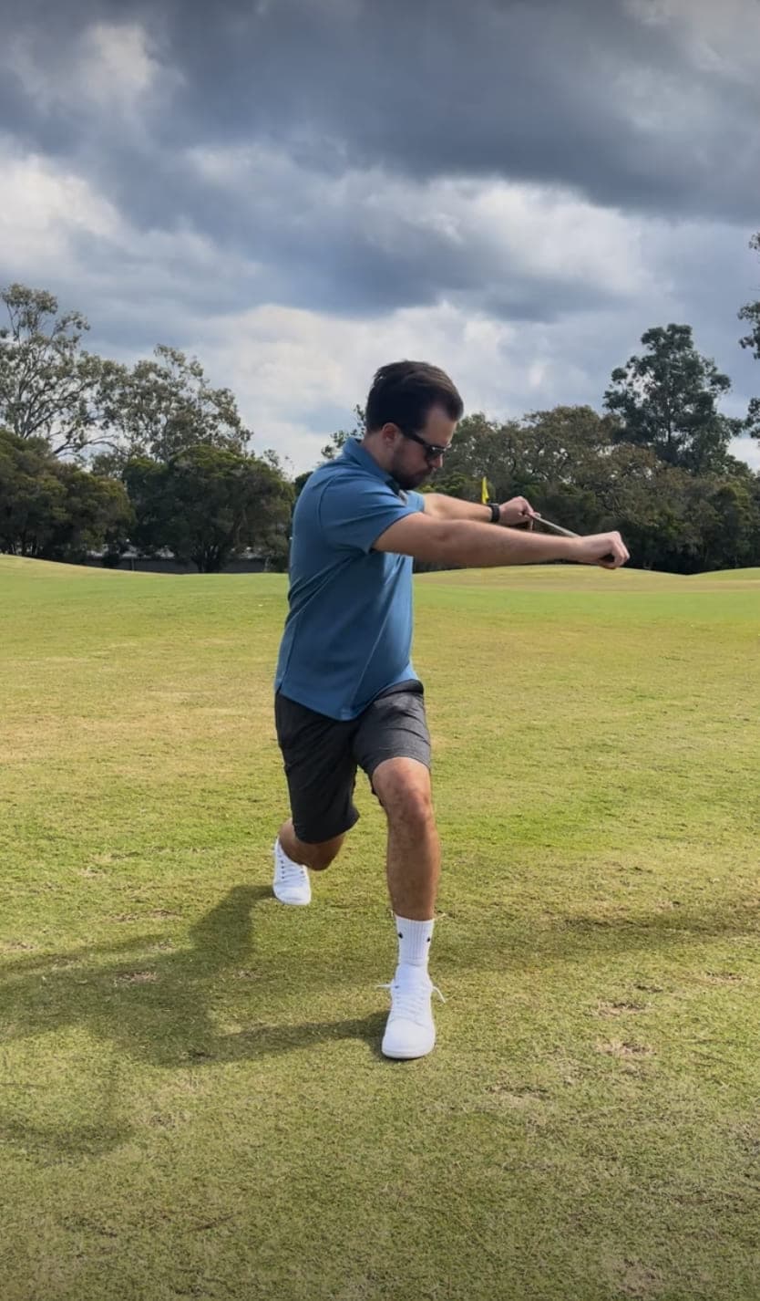 Cameron Radburn on a golf course in a reverse lunge, rotating through the torso with a golf club held out in front.