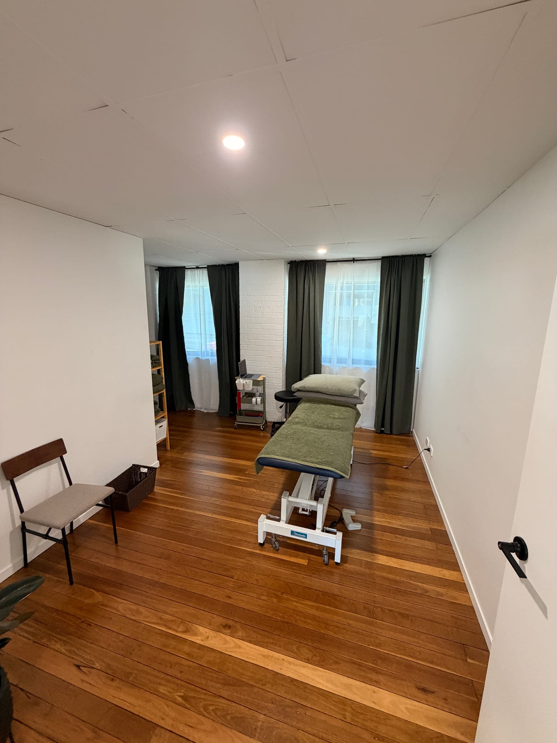 The TLP Physio treatment room, with a height-adjustable plinth, a chair for consultations, polished timber floors and natural light from the window.